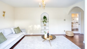 Een woonkamer met een witte bank, een ronde salontafel, een kroonluchter en een witte deur die naar een keuken leidt.