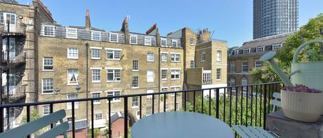 A balcony with a white table and chairs, overlooking a garden and a row of brick buildings.