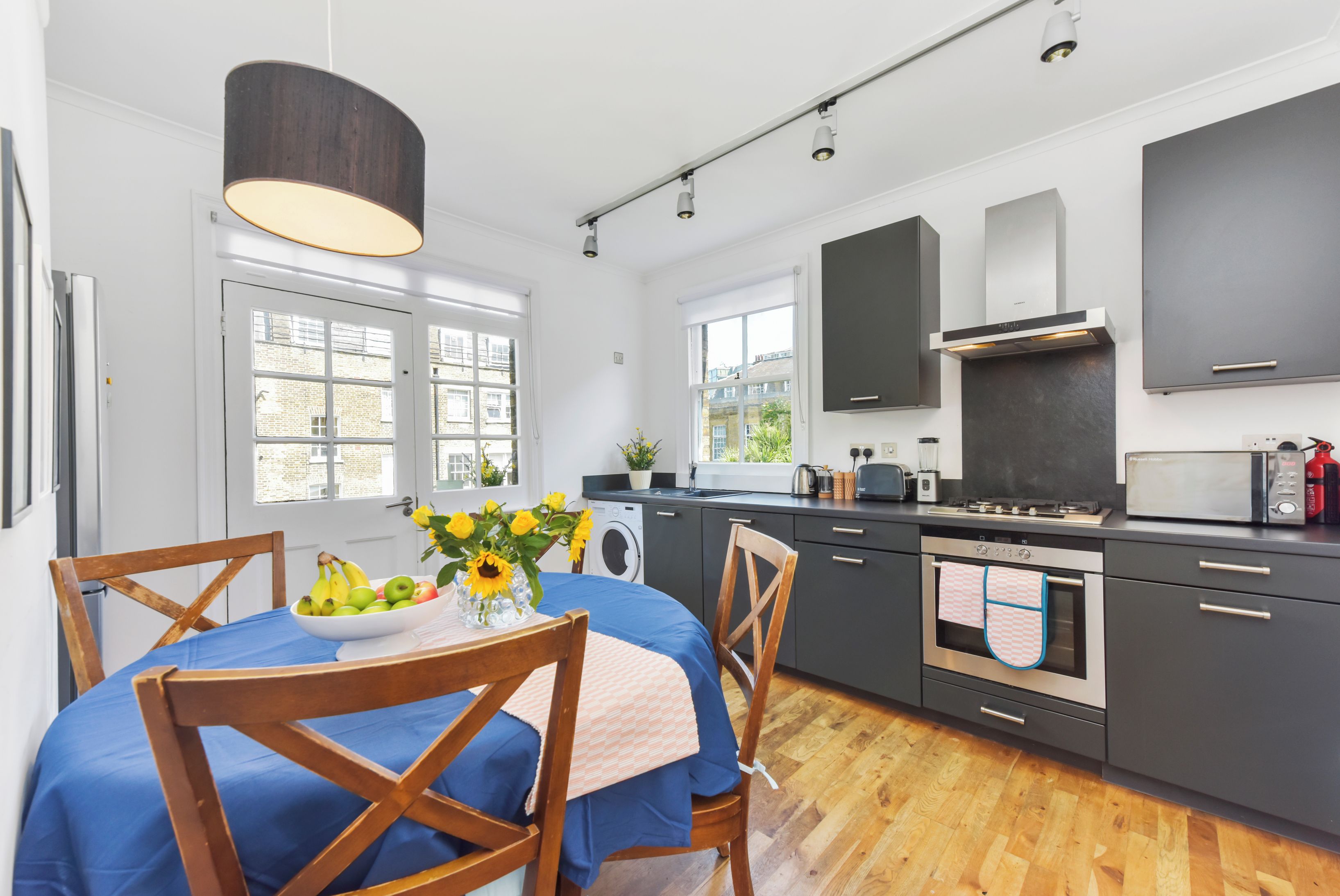 A modern kitchen with a dining area, featuring a wooden table with a blue tablecloth, a bowl of fruit, and a vase of flowers.