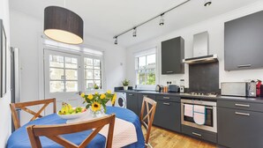 A modern kitchen with a dining area, featuring a wooden table with a blue tablecloth, a bowl of fruit, and a vase of flowers. - Spacious 3BDR On The Southbank With Balcony (Greater London)