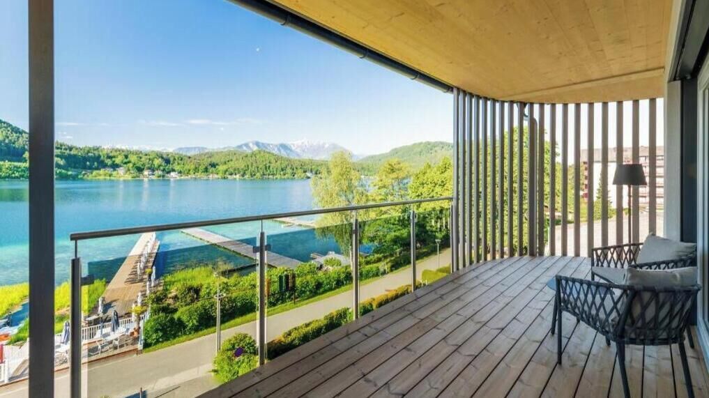 A balcony with a view of a lake and mountains, a wooden deck, and outdoor furniture.