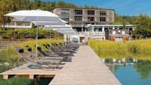 Une jetée en bois mène à une piscine agrémentée de chaises longues et de parasols, avec un bâtiment moderne en arrière-plan.
