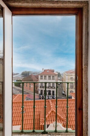 A view through a window with a balcony railing, a street with parked cars, and buildings with red-tiled roofs. - Bright Studio With Open Views Alfama (Lisbon)