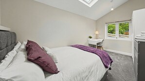 Ein Schlafzimmer mit Dachfenster, einem Bett mit weißer und violetter Bettwäsche, einem Schreibtisch mit Lampe und Blick auf Grünfläche vor dem Fenster.