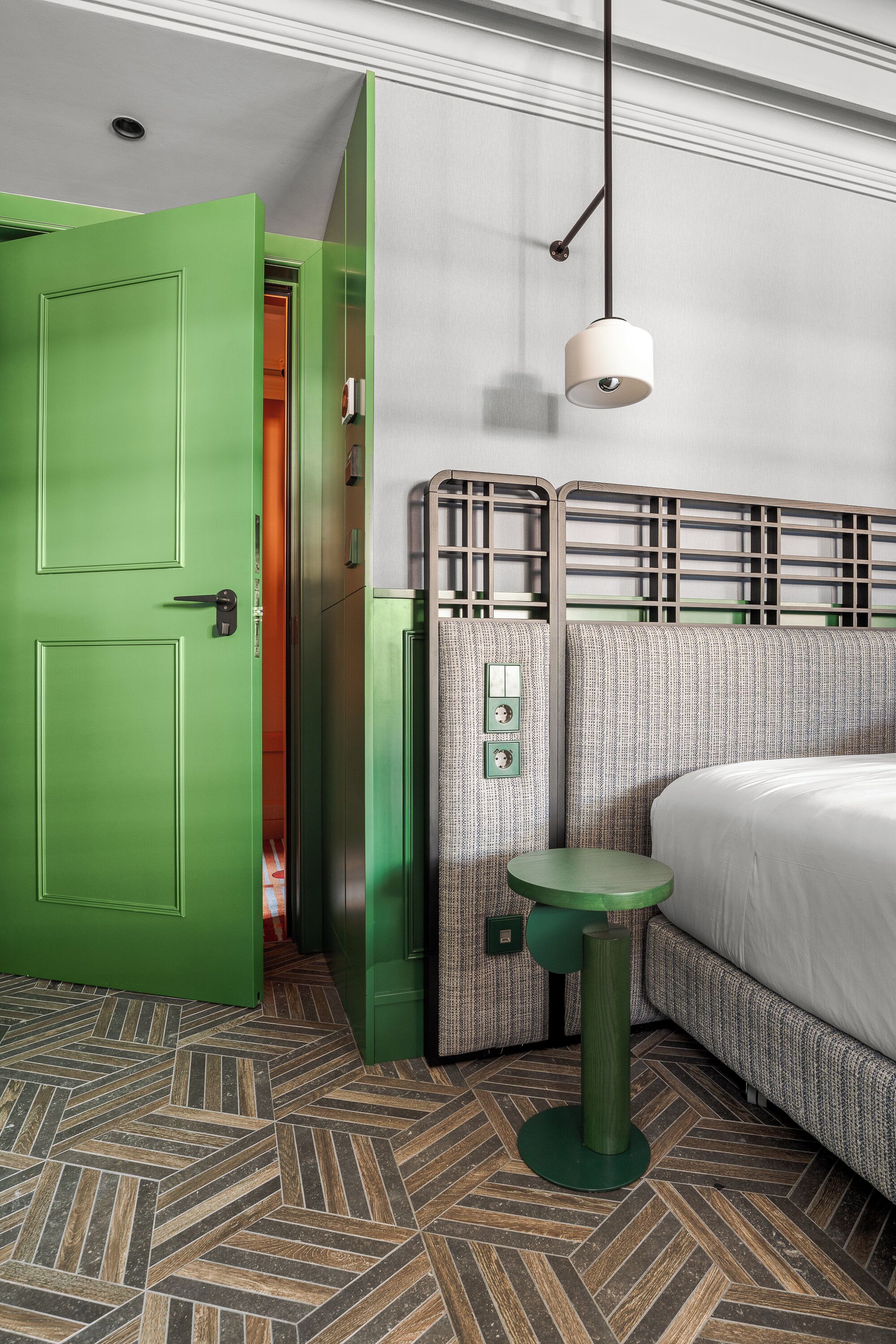 Habitación de hotel con puerta verde, cama con cabecera estampada, una mesita y una lámpara colgante.