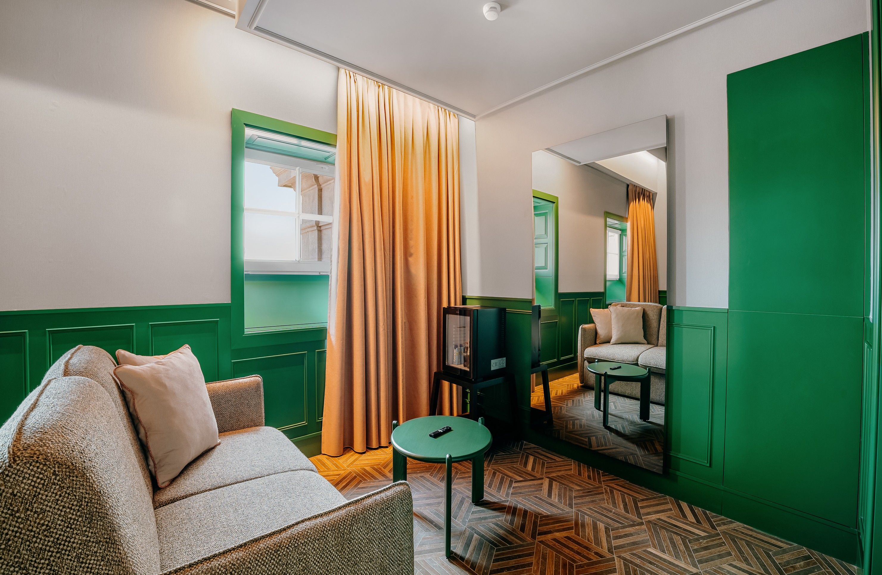 A hotel room with a green and white color scheme, a sofa, a small table, a television, and a mirror reflecting the room.