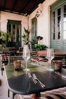 Mesa lista para cenar con dos copas de vino y cubiertos, rodeada de sillas de mimbre y una puerta verde al fondo.
