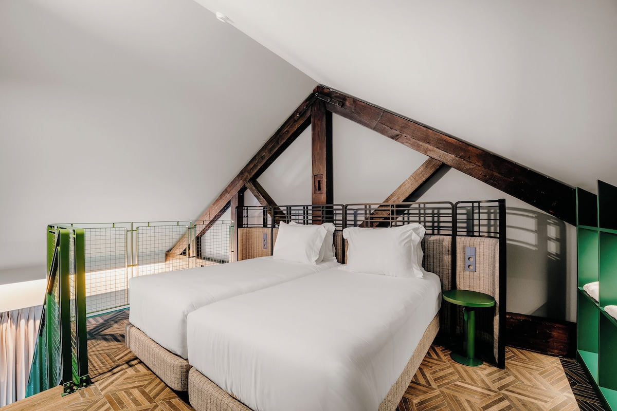 A bedroom with a high ceiling, exposed wooden beams, a bed with white linens, a small stool, and a green chair.