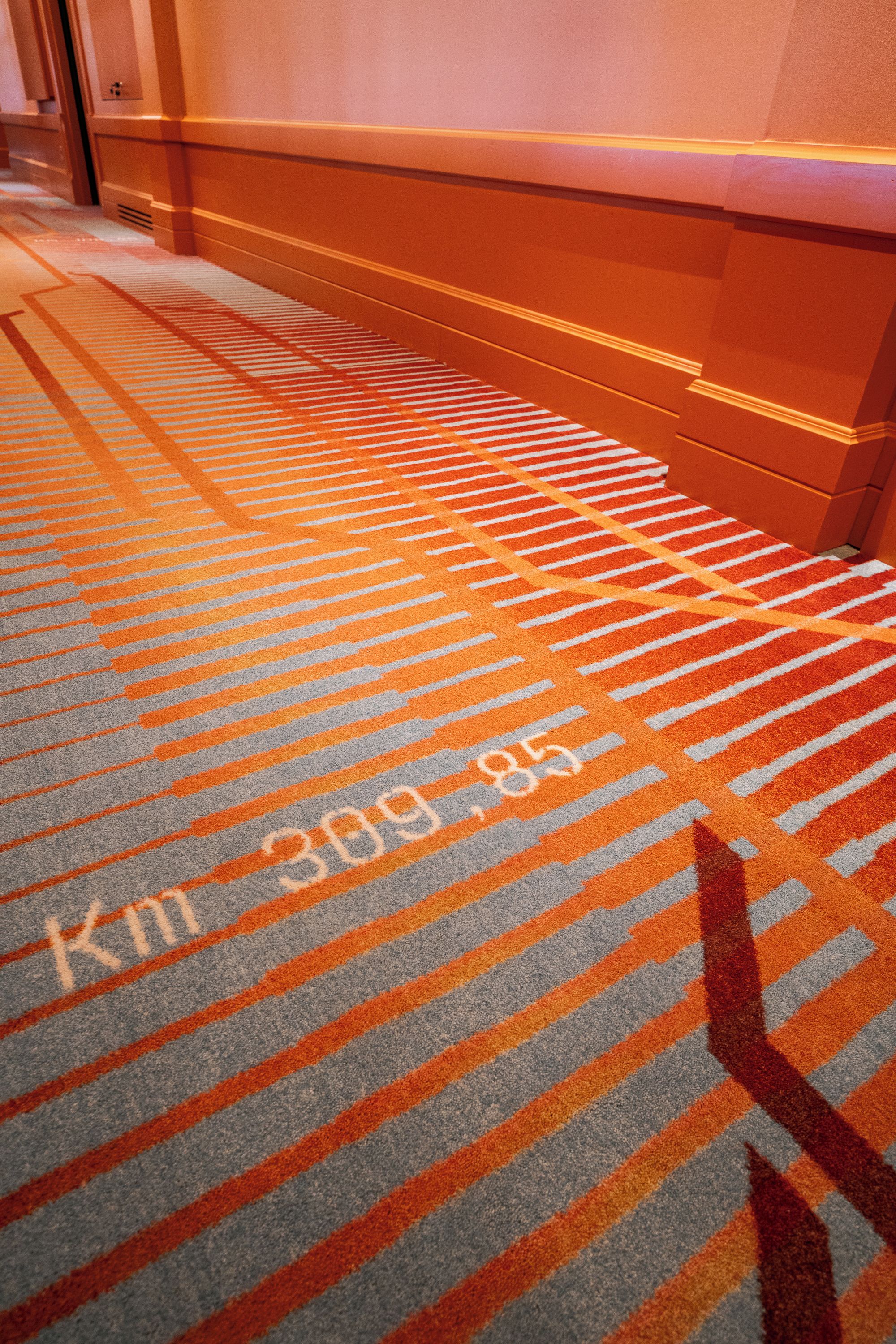 On peut voir un tapis orné d’un motif de rayures diagonales dans des tons d’orange et de gris, et l’inscription « km 309,85 » est visible.