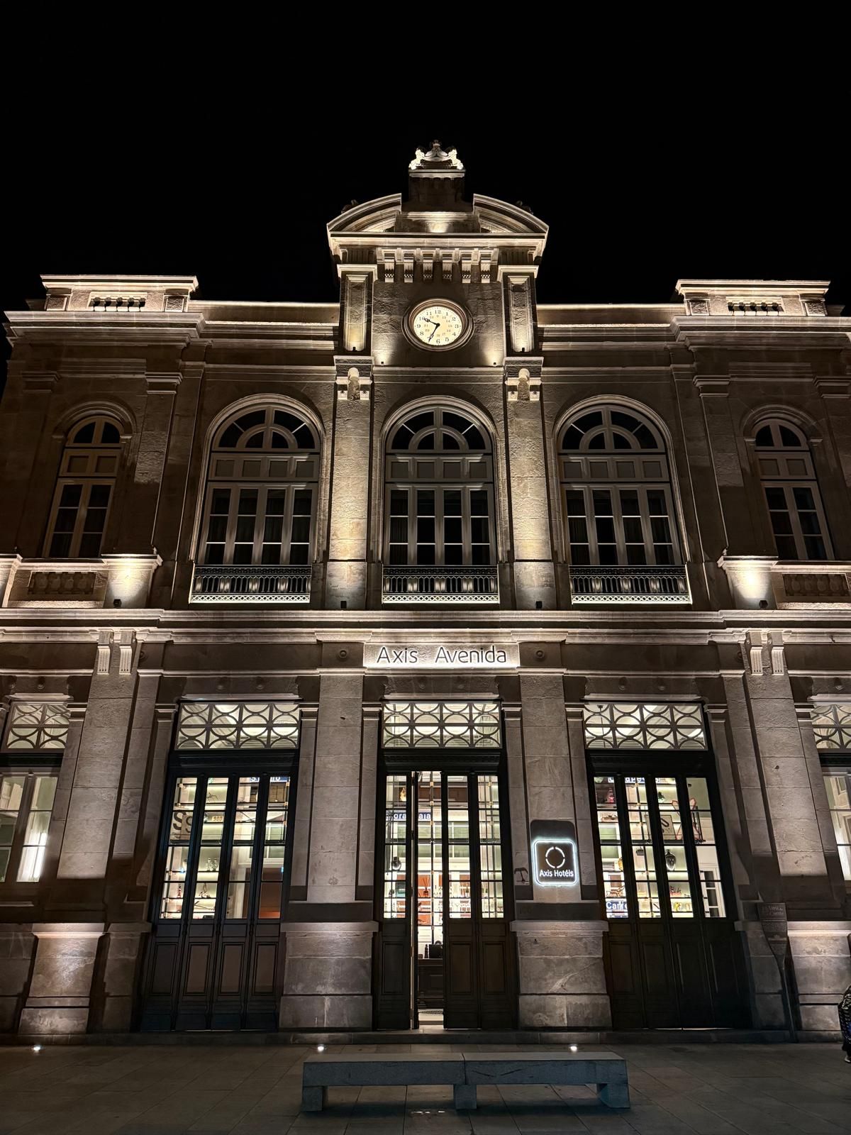 Un edificio imponente con una torre de reloj, amplias ventanas en arco y un letrero que dice 'Axis Avenida'.