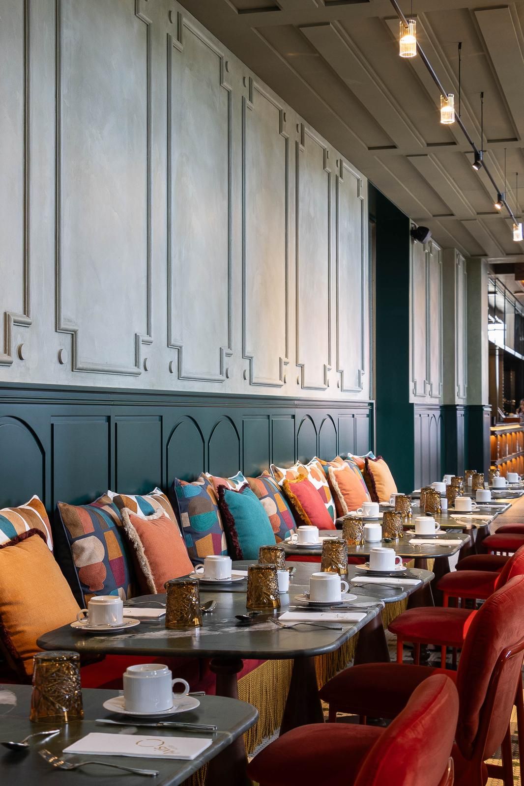 Une rangée de tables entourées de chaises, chacune agrémentée d’une tasse et d’une soucoupe blanches, ainsi que d’un coussin à motifs.