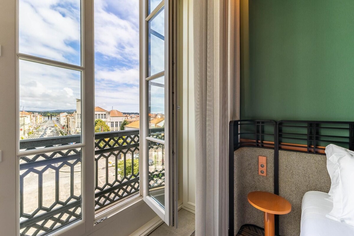 A hotel room with a view of a cityscape through large windows, a bed with a headboard, and a small table.