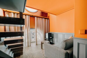 Una habitación moderna con suelo de espiga, un sofá gris, un televisor sobre un mueble bajo y una ventana con cortinas.