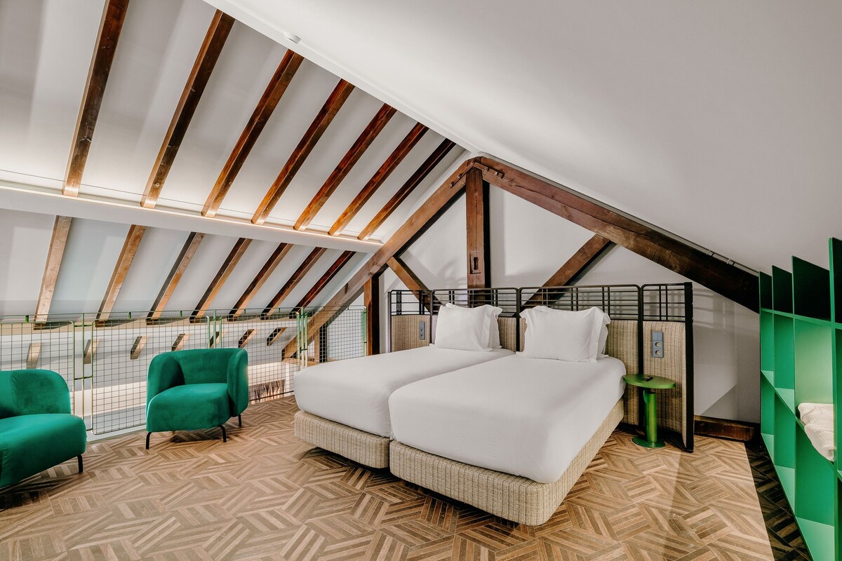 A bedroom with a sloped ceiling, a bed with white bedding, a green chair, and a small table.