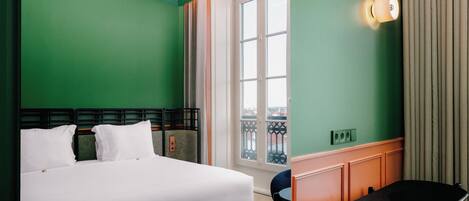 A hotel room with a bed, a chair, a window, and a wall-mounted light fixture.