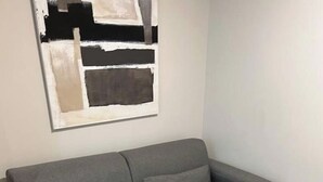 Un canapé gris avec deux coussins, une table ronde en bois avec un pied noir, et un tableau abstrait moderne accroché au mur.
