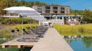 Une jetée en bois mène à une piscine bordée de chaises longues et d’ombrelles, avec un bâtiment visible au loin.