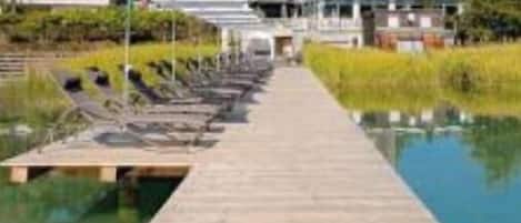 Un muelle de madera que da a una piscina con sillones de playa y sombrillas, con un edificio al fondo.