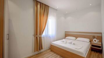 A modern hotel room with a wooden bed, white linens, a nightstand, and a window with sheer curtains.