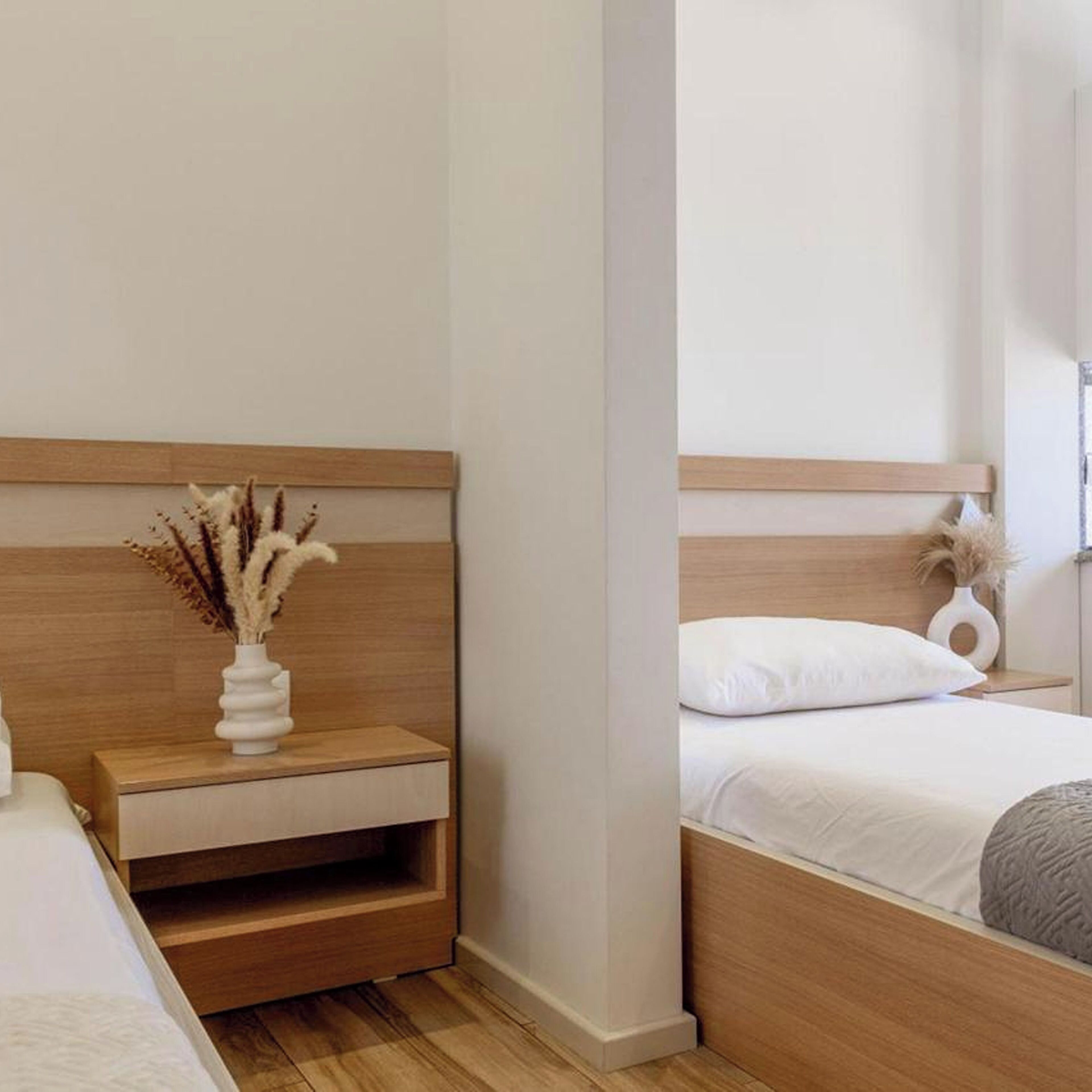 Une chambre d’hôtel avec une tête de lit en bois, une table de chevet ornée d’un vase contenant des plantes séchées, un lit avec une literie blanche et une table de nuit sur laquelle est posé un objet décoratif.