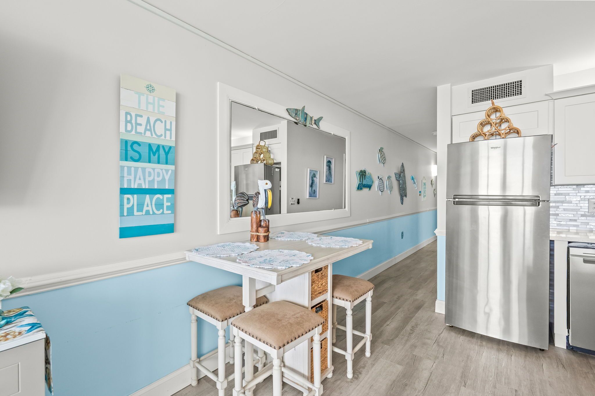 A kitchen with a stainless steel refrigerator, a white table with two chairs, and a wall with a beach-themed sign.