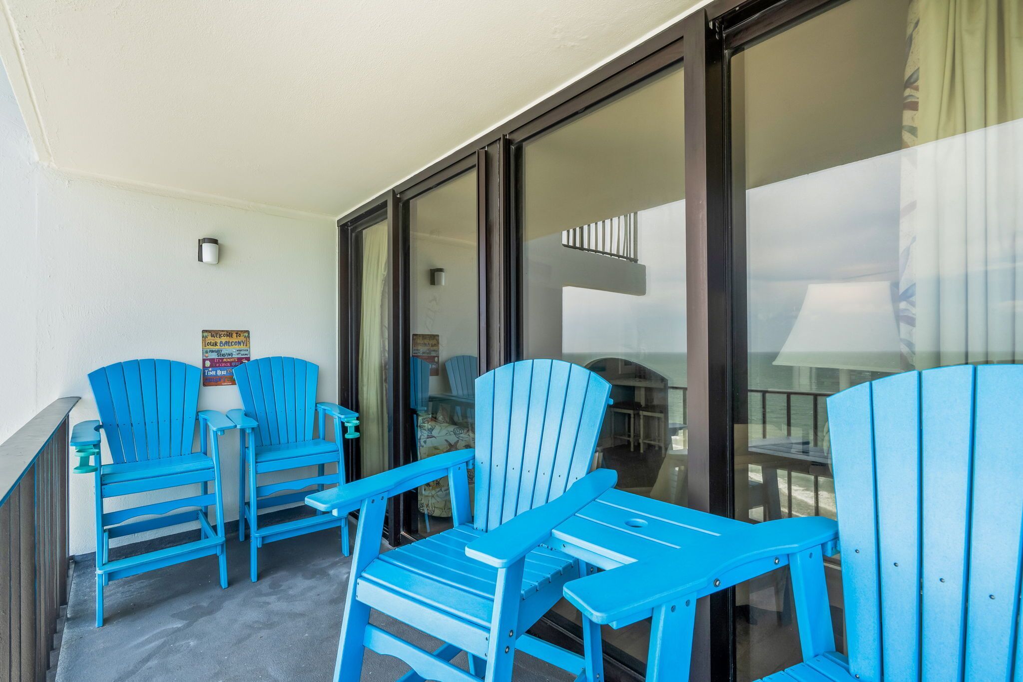 A balcony with three blue Adirondack chairs, a small table, and a sign welcoming guests to the balcony.