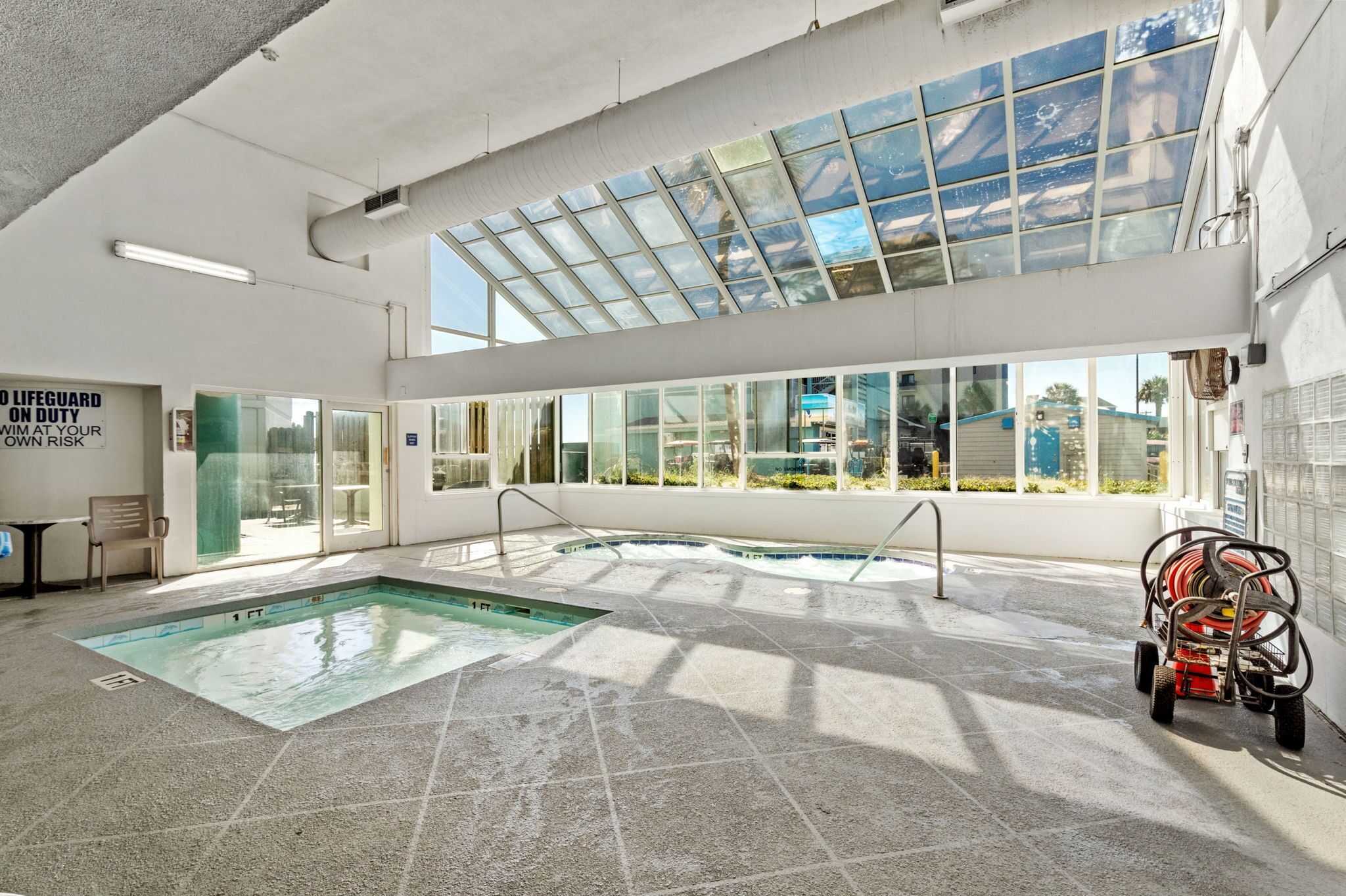 An indoor swimming pool with clear glass walls, a lifeguard sign, and a hose reel on the right.