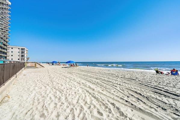 A beach with a high-rise building on the left, clear skies, and a sandy shore with footprints. - Royal Garden Resort 1214 (Murrells Inlet)