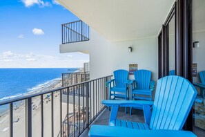 A balcony with a view of the beach and ocean, featuring blue chairs and a small table. - Royal Garden Resort 1214 (Murrells Inlet)