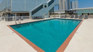 A swimming pool with a brick border, surrounded by a deck with chairs and tables.