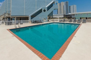 A swimming pool with a brick border, surrounded by a deck with chairs and tables.
