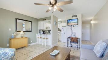 A living room with a wooden coffee table, a sofa, a ceiling fan, and a kitchen area with white cabinets and a TV.