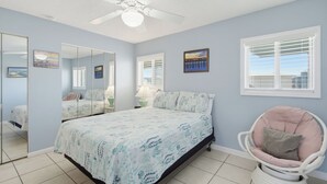 Een slaapkamer met een bed, een stoel, een plafondventilator, een raam met zonwering, en een muur met ingelijste foto's.