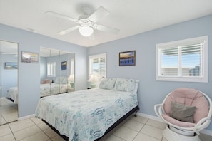A bedroom with a bed, a chair, a ceiling fan, a window with blinds, and a wall with framed pictures. - Summer House West B105 (Gulf Shores)