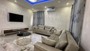 A living room with a flat-screen TV, beige sofa, and a coffee table. - Prime Residence Apartments - Dalaba (Old-Yundum)