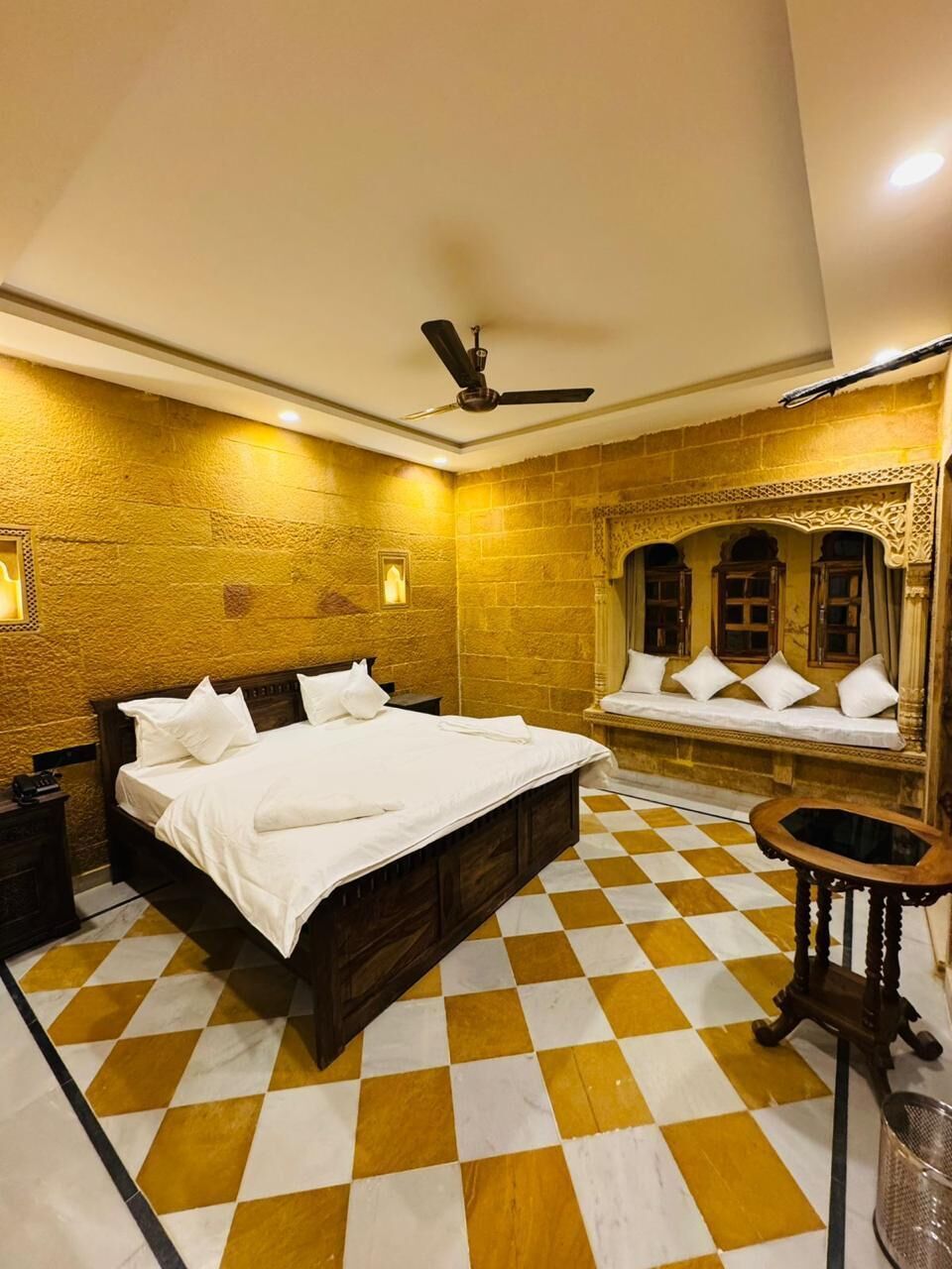 Habitación de hotel con piso a cuadros, una cama con ropa blanca, un ventilador de techo y un banco con cojines pegado a la pared.