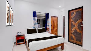 A bedroom with a bed, a nightstand, a window with curtains, a wooden door, and a wall-mounted artwork. - Deluxe Room Ravai (Calangute)
