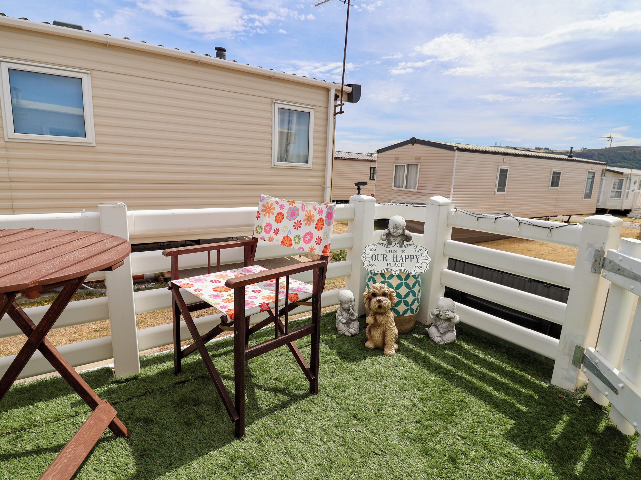 Un espace patio avec une table et des chaises en bois, une clôture décorative et un panneau indiquant : « ICI, C’EST NOTRE PETIT COIN DE PARADIS. »