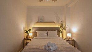 A neatly made bed with a folded towel, two bedside lamps, a framed picture above the bed, and a ceiling fan. - Apartment Quart Mid-stay (Valencia)