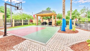 Ein Basketballplatz im Freien mit einer Laube, Palmen und einem gepflasterten Weg.