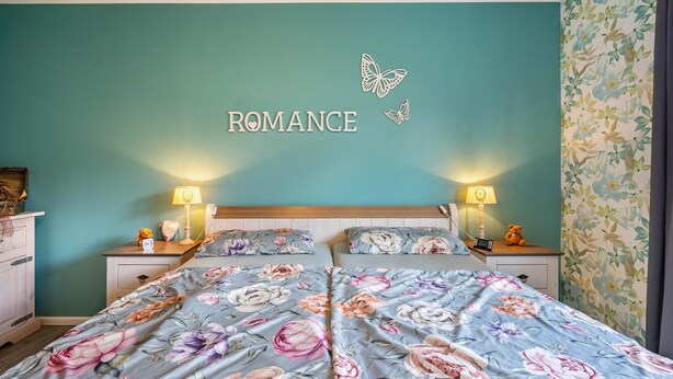 Une chambre avec un mur de couleur turquoise, une literie à motifs floraux, deux tables de chevet avec des lampes et un papillon décoratif.