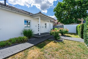 A single-story house with a white exterior, a front porch, and a small garden with a rose bush.