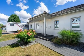 Ein eingeschossiges Gebäude eines Wohnmobilparks mit überdachter Veranda, einem kleinen Garten mit Blumen und einem abgestellten Wohnmobil.