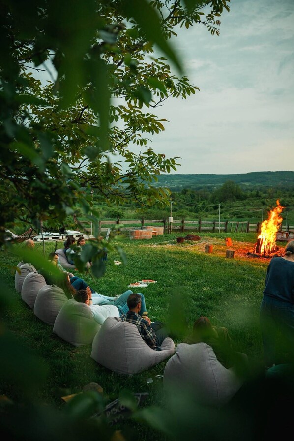 People sitting on bean bags around a campfire in a grassy outdoor area with trees and hills in the background.