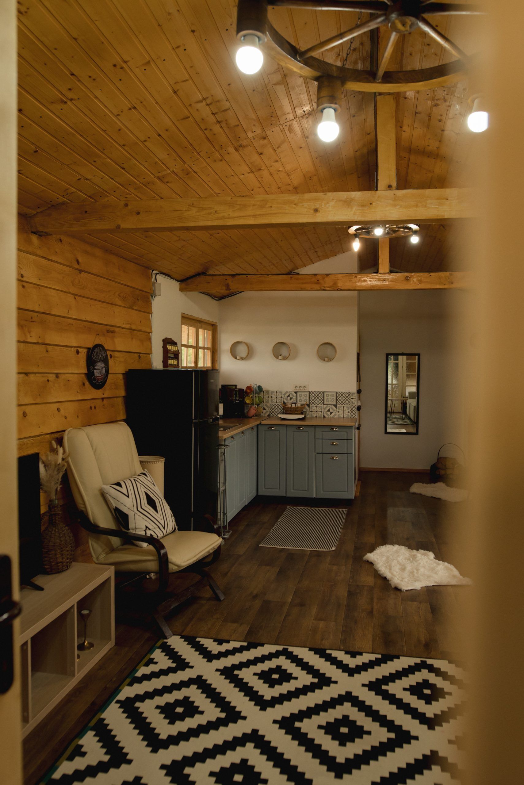 Un interior acogedor con techo de madera, una zona de cocina con armarios y un refrigerador, y un comedor con una silla y una alfombra.