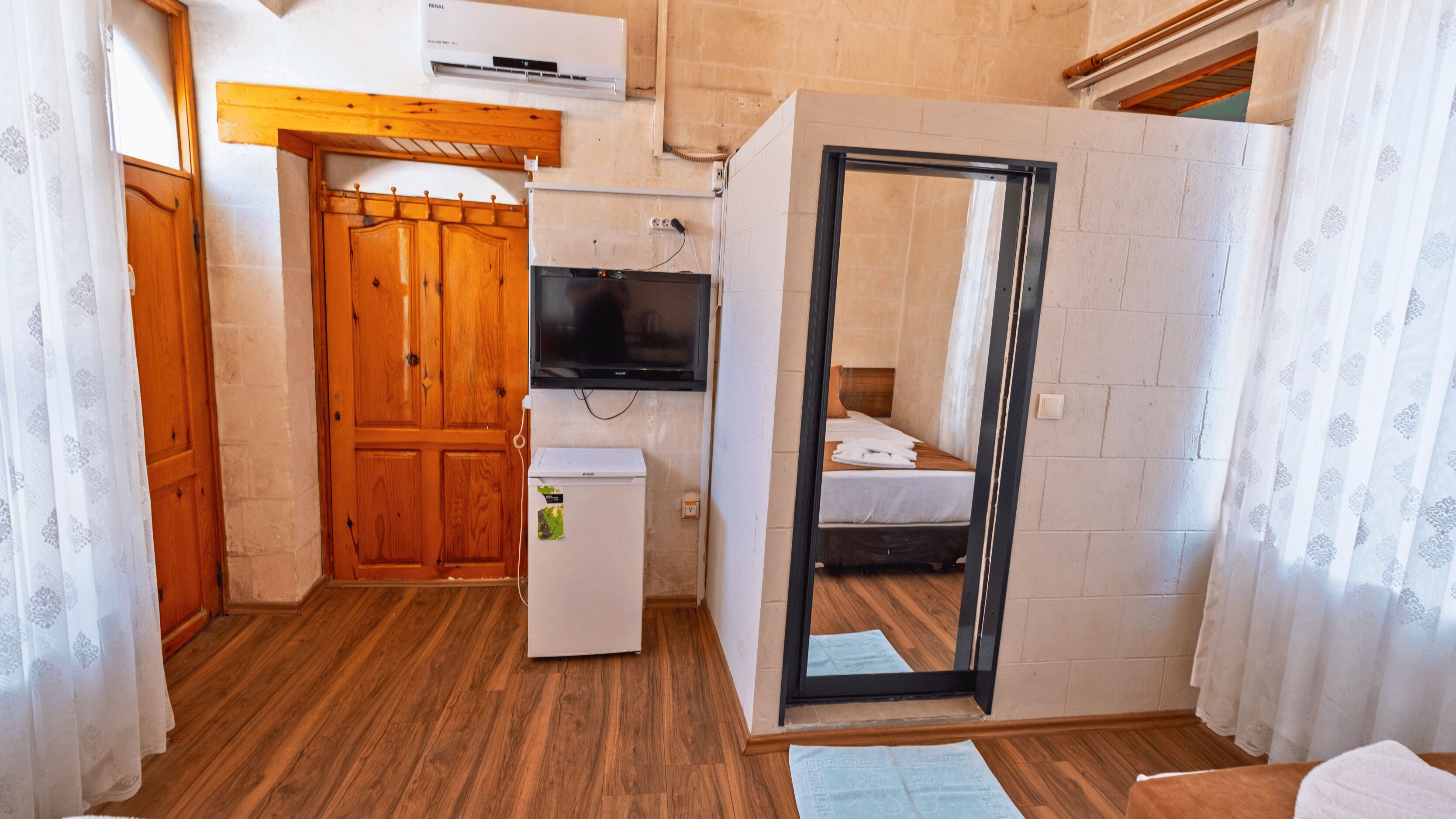 Habitación compacta con puerta de madera, un refrigerador pequeño blanco, un televisor de pantalla plana montado en la pared y una cama con cubrecama blanco.