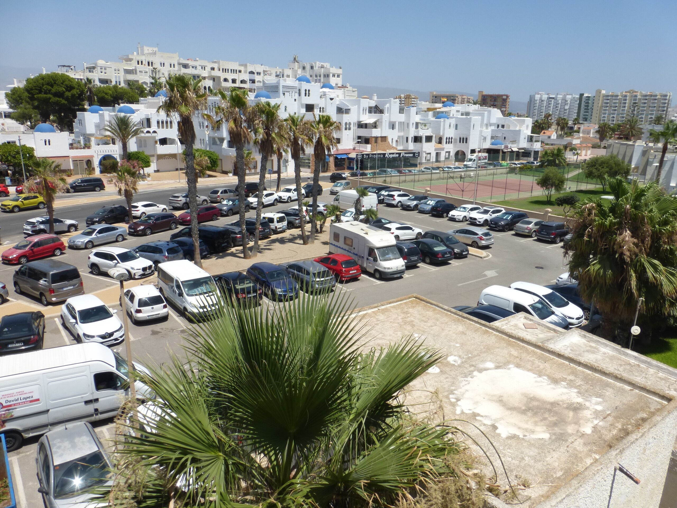 Un estacionamiento con varios coches y algunas camionetas, rodeado de palmeras y edificios de fachadas blancas y cúpulas azules.