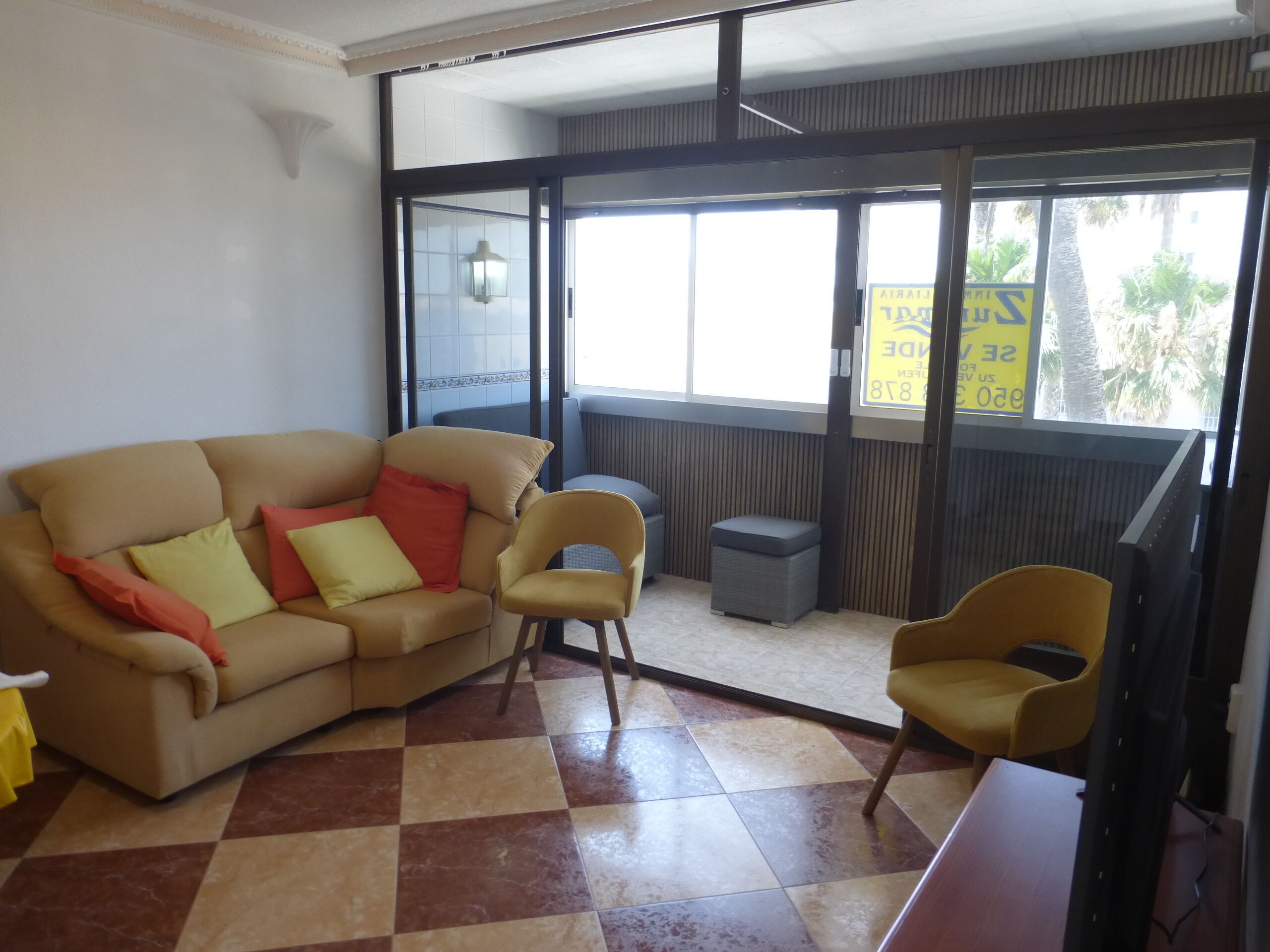 Een kamer met een beige bank, twee fauteuils, een bijzettafeltje en een raam met uitzicht op palmbomen.