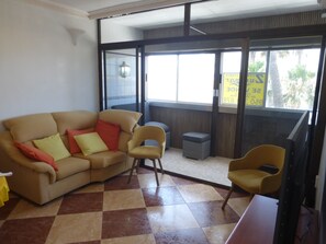 Ein Zimmer mit beiger Couch, zwei Sesseln, einem kleinen Tisch und einem Fenster mit Blick auf Palmen.