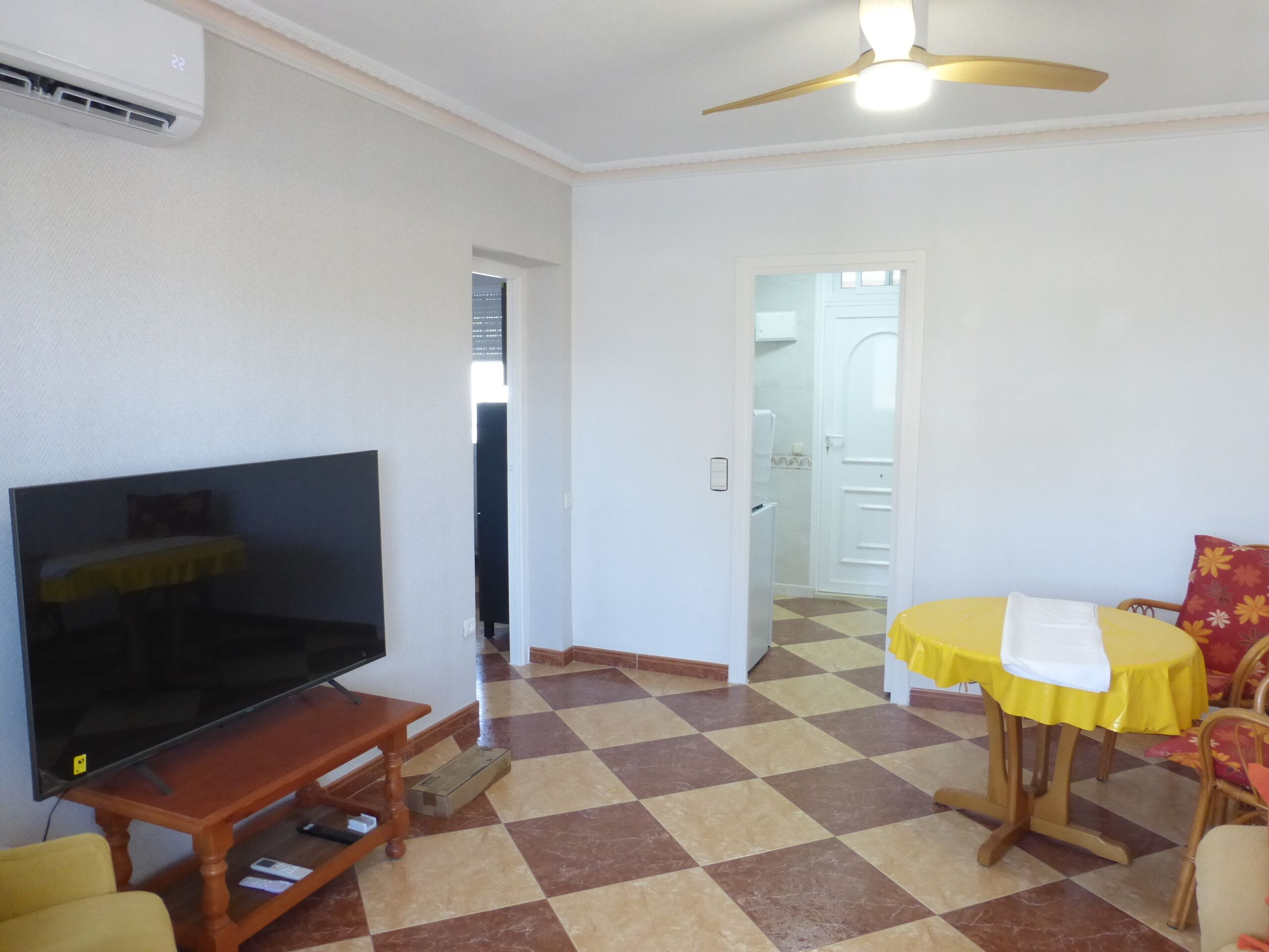 Una sala de estar con un televisor de pantalla plana sobre un soporte de madera, un comedor con una mesa y sillas amarillas, y un piso a cuadros.